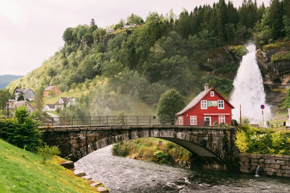 Rotes Haus hinter Brücke, neben Fluss vor Wasserfall im sommerlichen Norwegen