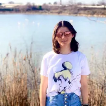 Austauschschülerin in T-Shirt und Sonnenbrille vor Seeufer mit Schilf bei Sonnenschein